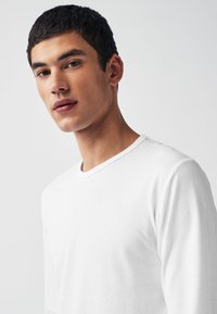 Vit långärmad t-shirt med rund halsringning, slät textur och normal passform, uppvisad av en modell mot en neutral bakgrund.