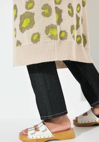 Knit-sweater beige väri, millel on rohelised ja kollased leopardilaigud, koos tumesiniste teksade ja valgete sandaalidega, millel on puidust platvorm ja hõbedased aktsendid.
