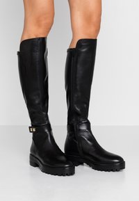 Bottes en cuir noir montant jusqu'au genou, au design épuré, dotées d'une semelle plate, d'une fermeture éclair latérale et d'un ornement métallique sur la bride à la cheville.