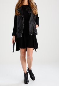 Svart läderväst utan ärmar med silverdetaljer över en svart knälång klänning, parat med svarta ankelskor. Slät textur och figurnära design.