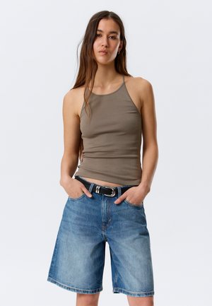Vrouw met lang bruin haar, een mouwloos taupekleurig topje en losse knielange spijkershorts met een zwarte riem. Handen in de zakken.
