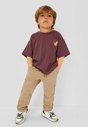 Young child with light brown hair wearing a brown shirt with a small pineapple patch, beige pants, and white sneakers standing against a plain background.