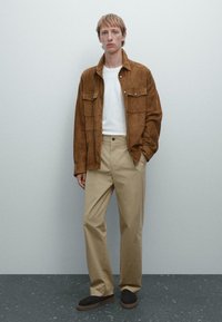 Brown suede jacket with two chest pockets over a white shirt; beige trousers; dark shoes; modern cut, smooth texture, and neutral colors.