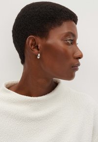 Profilo di una persona con capelli corti e ricci, che indossa una maglietta bianca texturizzata e un piccolo orecchino a forma di goccia in argento.