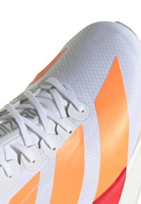 Primer plano de una zapatilla atlética blanca con cordones blancos y llamativas rayas naranjas sobre una superficie superior texturizada.