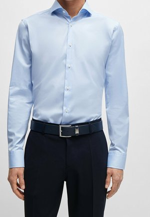 Mężczyzna w jasnoniebieskiej koszuli z długim rękawem, wpuszczonej w ciemne spodnie z czarnym paskiem, na jednolitym jasnym tle.