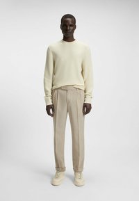 Maglione beige chiaro con texture, pantaloni chiari su misura e sneakers color crema. Modello in piedi contro uno sfondo neutro.