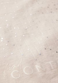 Ljust beige tyg med spridda silverpaljetter och präglad text som säger "find CON." Slät textur med subtila mönster.