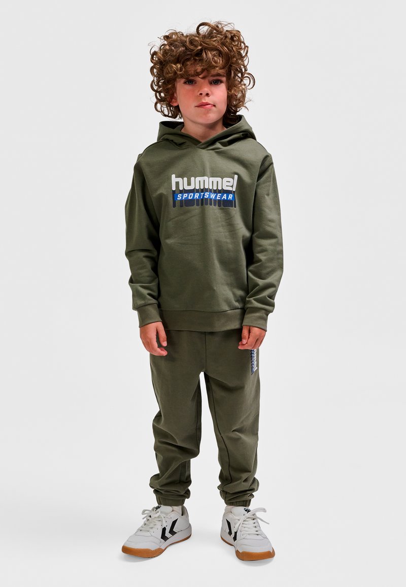 Jeune enfant aux cheveux bouclés portant un sweat à capuche de sport hummel vert olive et un pantalon de jogging assorti avec des baskets blanches sur fond blanc.