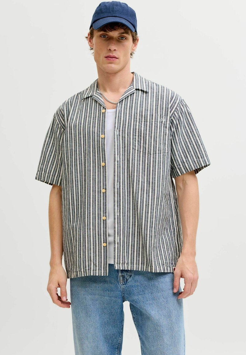 Jonge man die een marineblauwe pet draagt, een grijs-wit verticaal gestreept shirt met korte mouwen over een wit T-shirt, en een blauwe spijkerbroek.