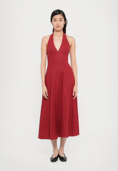 Une longue robe à col halter en tissu rouge foncé avec un corsage ajusté, une jupe évasée et une texture subtile, associée à des ballerines noires.
