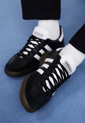 Baskets noires Adidas Handball Spezial avec des bandes et des lacets blancs, portées avec des chaussettes blanches et un pantalon noir sur une surface bleue.