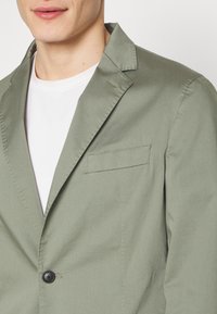 Grüner Baumwollblazer mit Reverskragen, einfacher Knopfleiste und einer Brusttasche. Glatte Textur mit dezenten Nähten.