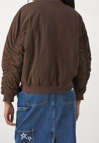 Henkilö, jolla on päällään ruskea, rypytetty hiha takki ja siniset farkkuhameet, joiden takataskussa on valkoisia tähtikuvioita, seisoo yksinkertaista taustaa vasten.