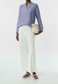 Blouse longue lilas à manches longues avec un col à encoches, pantalon blanc à jambes larges et sandales plates blanches. Petit sac à main blanc de forme arrondie.
