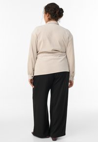 Bež srajca z dolgimi rokavi in ovratnikom ter pasom, v kombinaciji s širokimi črnimi hlačami. Mehka tkanina z diskretno teksturo in gumbi.