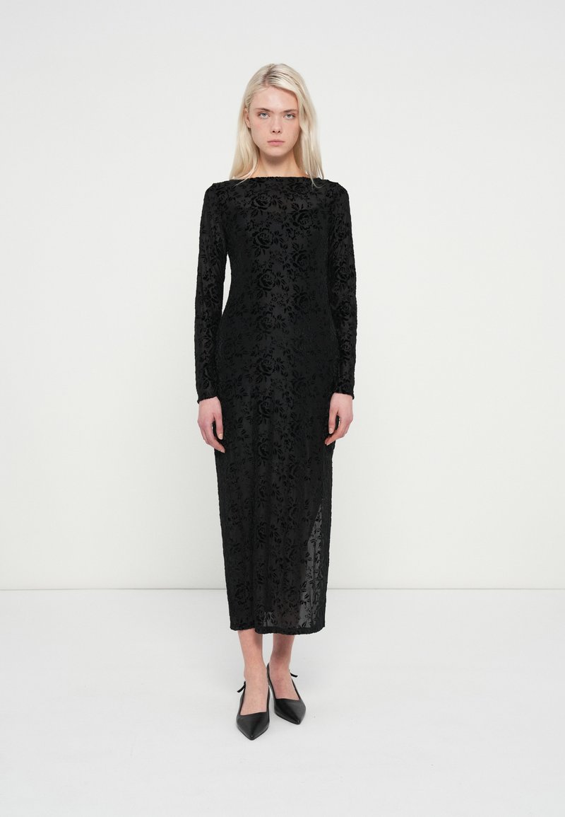 Μαύρο φόρεμα μακρυμάνικο με λουλουδάτο δαντελένιο μοτίβο, ίσια σιλουέτα και ένα πλαϊνό σκίσιμο, σε συνδυασμό με μυτερές μαύρες γόβες.