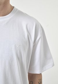 Weißes Baumwoll-T-Shirt mit Rundhalsausschnitt, kurzen Ärmeln und einem kleinen gestickten Logo auf der linken Brust. Glatte Textur, lässige Passform.