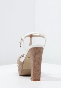 Brun plattformssandal med vävd textur, vit ankelrem och hög blockklack. Metallspänne som accent på remmen.
