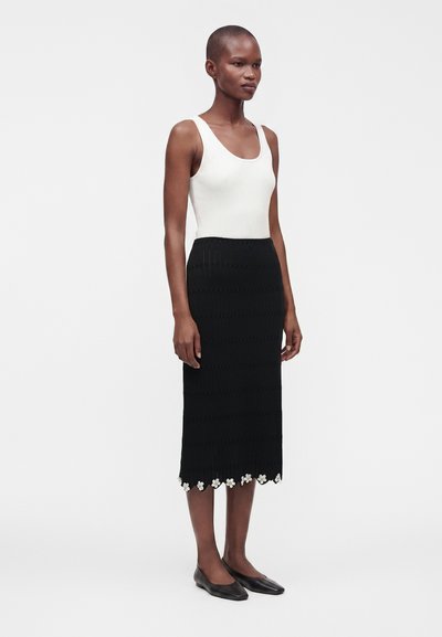 Femme portant un haut sans manches blanc et une jupe midi noire avec un bord floral blanc à l'ourlet, debout devant un fond uni.