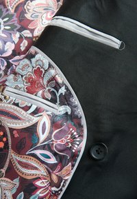 Schwarzer Stoff mit einem floralen Paisleymuster in Burgunderrot, Lila und Türkis, mit einem Knopf und weißen Paspeln an der Tasche.