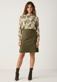 Blouse florale en vert et rose avec des détails volantés, associée à une jupe en daim vert olive ajustée avec des poches latérales et des zips.