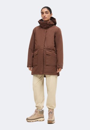 Person iført en brun hooded vinterjakke, beige joggebukser og beige vandrestøvler, stående mod en ensfarvet lys baggrund.