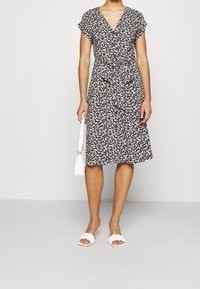 Donna che indossa un vestito nero a fiori lungo fino al ginocchio con bottone frontale e cintura, tiene in mano una borsa bianca e indossa sandali con tacco intrecciati bianchi.