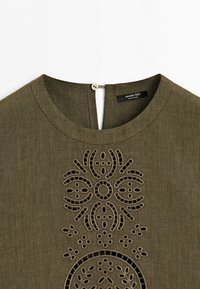 Olive green linen dress with round neck and button closure, featuring intricate cut-out embroidery patterns on the chest.