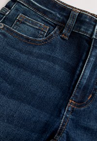 Primo piano di jeans in denim blu scuro che mostrano la tasca anteriore, la tasca piccola per le monete, il passante per la cintura e i dettagli di cuciture arancioni.