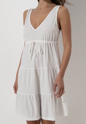 Donna che indossa un vestito bianco senza maniche a balze, con scollo a V e vita con coulisse, in piedi davanti a uno sfondo neutro.