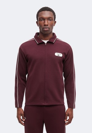 Uomo che indossa una giacca bordeaux con zip e pantaloni coordinati con piping bianco su maniche e gambe, su sfondo uniforme.