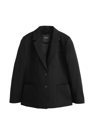 Junge Frau mit lockigem Haar, die einen schwarzen Blazer über einem weißen Hemd und einem schwarzen Rock trägt, steht vor einem einfachen hellen Hintergrund.