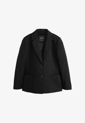 Ung kvinde med krøllet hår iført en sort blazer over en hvid skjorte og et sort nederdel, stående mod en ensartet lys baggrund.