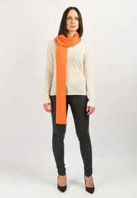 Orange uld tørklæde svøbt om halsen, parret med en lys sweater og mørke skinny jeans. Tørklædet har en blød tekstur og lang form.