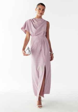 Vestido largo lavanda con hombro asimétrico y profunda abertura lateral. Tejido liso, ajuste entallado, combinado con un bolso de mano plateado y tacones blancos.