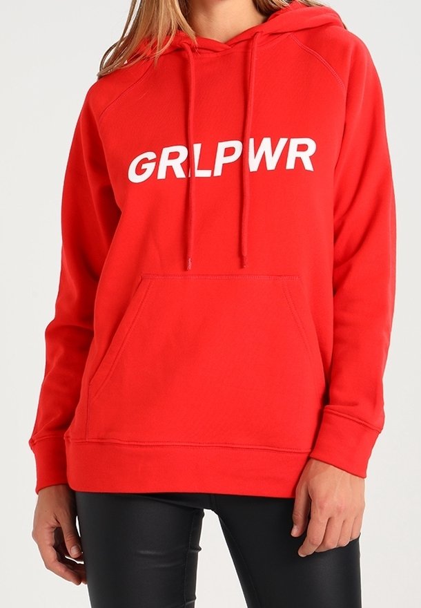 Femme portant un sweat à capuche rouge avec un texte blanc "GRLPWR" et un pantalon noir, debout devant un fond uni.