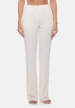 Donna che indossa pantaloni bianchi a vita alta, ampi con piega frontale, in piedi a piedi nudi su sfondo bianco, mani rilassate lungo i fianchi.