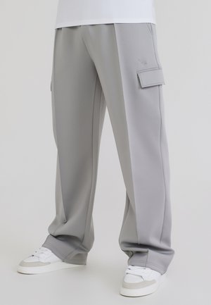 Pantalones cargo gris claro con bolsillos laterales, usados con zapatillas blancas y grises sobre un fondo liso.