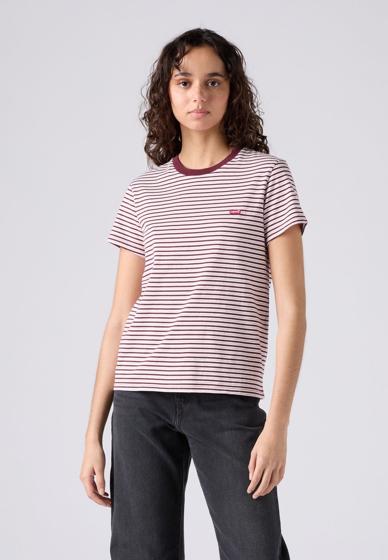 Jeune femme aux cheveux bouclés portant un t-shirt Levi's blanc à rayures bordeaux et un jean noir, debout devant un fond gris uni.