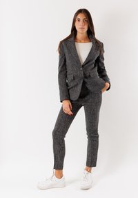 Completo grigio strutturato con giacca su misura e pantaloni aderenti, abbinato a scarpe da ginnastica bianche. Presenta una chiusura a un bottone e tasche.
