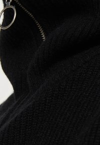 Schwarzer Strickpullover mit hohem Kragen und Reißverschluss, strukturiertem Gewebe und einem auffälligen silbernen Ringgriff am Reißverschluss.