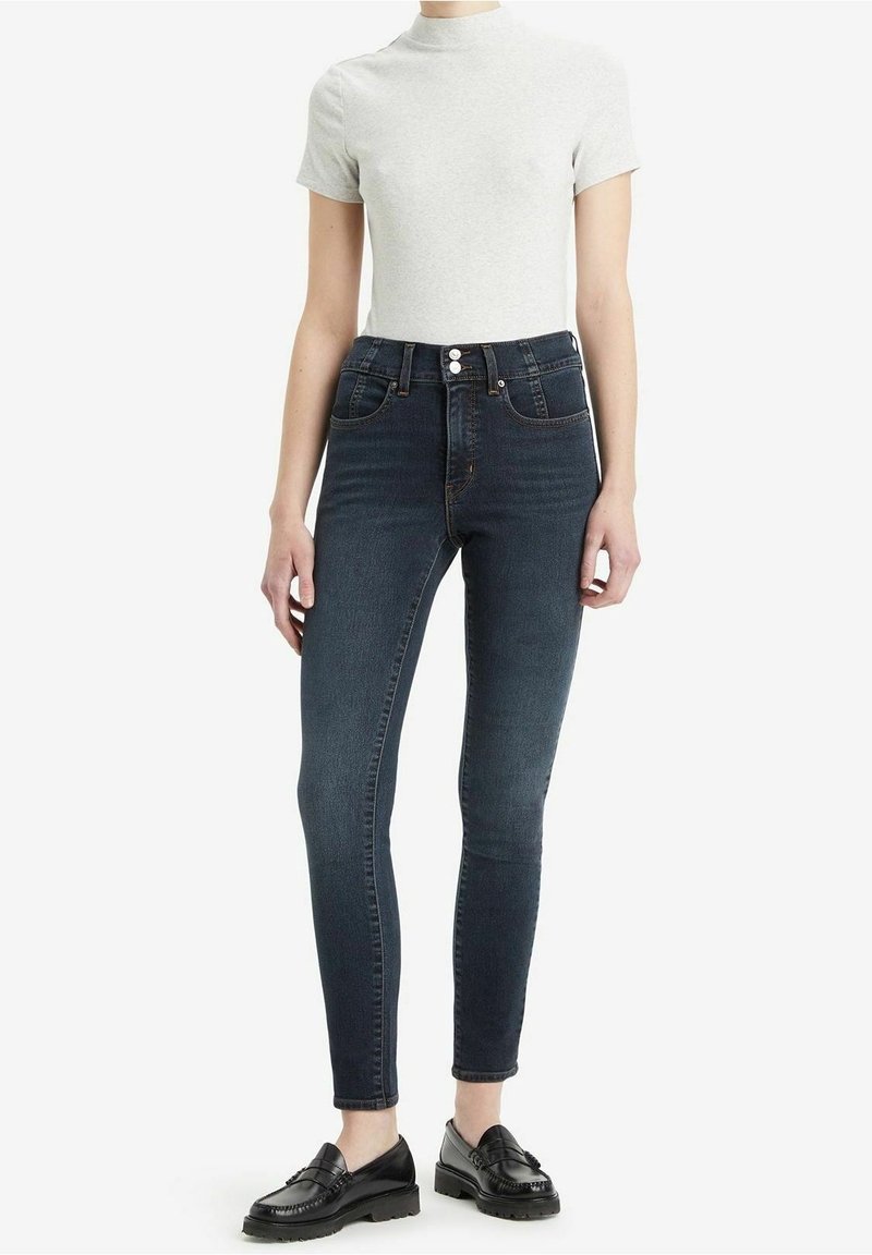 Jean skinny en denim foncé avec taille haute, cinq poches et fermeture à deux boutons. Associé à un haut ajusté gris clair à manches courtes et des mocassins noirs.