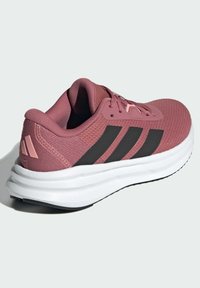 Pink athletic shoe with a textured mesh upper, three black stripes on the side, and a white cushioned sole. Lightweight design.