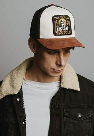 Mladenič z črno-belo tovornjakarsko kapo z rjavim šiltom in izvezeno značko z napisom "BLACKSKIES Saint of Sin Grace is Gone."