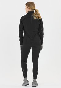 Svart sportjacka med hög krage och reflektande accenter, tillsammans med matchande svarta leggings som har ett randigt mönster och fickor med dragkedjor.