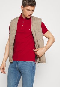 Lettvekts beige ermeløs vest med quiltet tekstur og sidelommer, båret over en tettsittende rød polo skjorte, sammen med blå jeans.