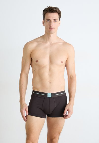 Bărbat fără tricou cu păr scurt, brunet, purtând boxeri negri cu o curea de talie gri și albastră, stând în fața unui fundal alb simplu.