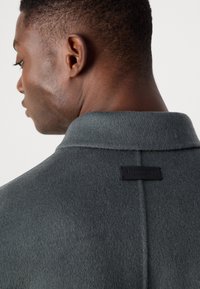 Gray wool blend coat with a soft texture, featuring a structured collar and a black brand label on the back. Minimalist design.