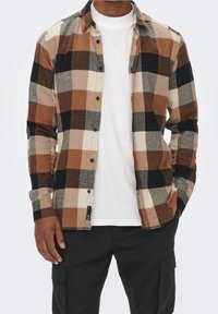 Camisa de franela a cuadros marrón y negro con mangas largas, cierre de botones y textura acolchada, combinada con una camiseta blanca debajo.